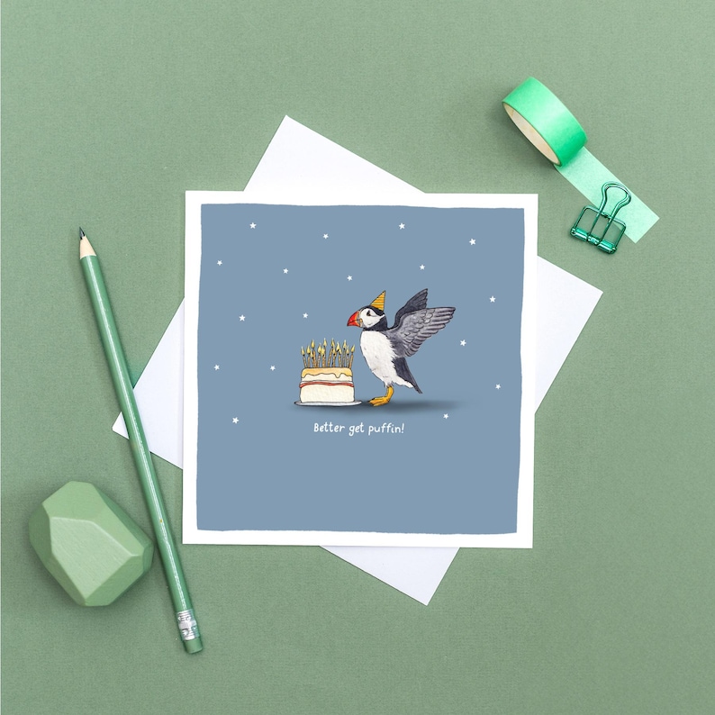 Pu&ograve; includere: Biglietto di auguri quadrato con un pulcinella di mare dei cartoni animati con un cappello da festa, accanto a una torta con candele accese. Il pulcinella di mare ha un'ala tesa. Il testo "Better get puffin!" &egrave; sotto la torta. Lo sfondo &egrave; blu con stelle bianche.