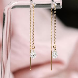 Puede incluir: Un par de pendientes de hilo de oro con cadenas finas y delicadas que suspenden una piedra preciosa transparente con facetas en forma de pera en cada extremo. Los pendientes cuelgan verticalmente de un soporte transparente.