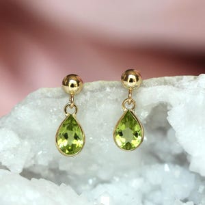 May include: A pair of gold stud earrings featuring dangling pear-shaped peridot gemstones. Each earring consists of a small polished gold sphere at the top, connected by a thin gold loop to a faceted, light green teardrop-shaped peridot stone.