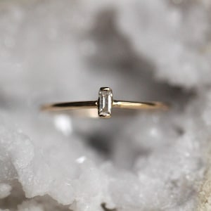 May include: A gold band ring with a single, clear, rectangular-shaped diamond set in the center.