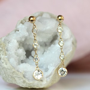 May include: A pair of gold drop earrings. Each earring has a small gold ball at the top, linked by a fine chain to a series of clear, round gemstones. The earrings are set against a white, textured background with a soft pink backdrop.