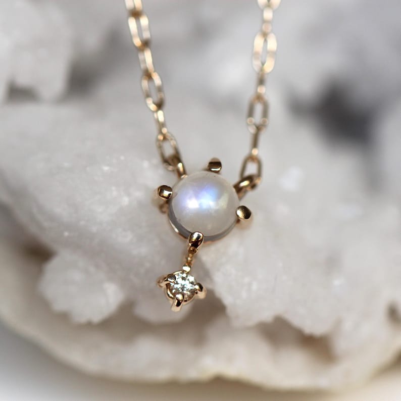 May include: A delicate gold necklace featuring a round moonstone pendant with a small diamond accent. The necklace is displayed against a white background.