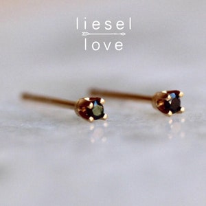 May include: A pair of gold stud earrings featuring small, round, dark-colored gemstones set in four-prong settings. The text "liesel love" with an arrow graphic is positioned above the earrings. The earrings have thin, straight posts.
