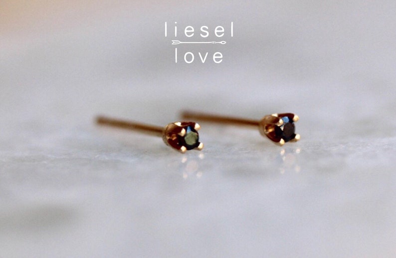 May include: A pair of gold stud earrings featuring small, round, dark-colored gemstones set in four-prong settings. The text "liesel love" with an arrow graphic is positioned above the earrings. The earrings have thin, straight posts.