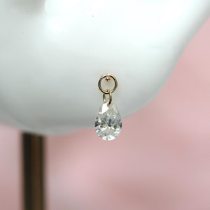 May include: A gold-toned earring featuring a small circular hoop attached to a clear, faceted, teardrop-shaped crystal pendant. The jewelry piece is displayed on a white stand, showcasing the reflective quality of the clear stone and the metallic finish of the circular hoop component.