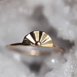 May include: A gold ring featuring a fan-shaped centerpiece with a smooth, polished finish. The design includes a central circular bead at the base of the fan, with radiating lines extending outward to the curved top edge of the decorative element.