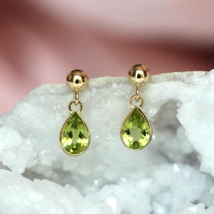 May include: A pair of gold drop earrings featuring faceted, pear-shaped light green peridot gemstones suspended from polished gold ball studs. The earrings are displayed against a textured, crystalline white mineral surface.