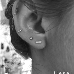 May include: A black and white image showing a set of silver-toned earrings worn in an ear. The jewelry includes a small stud, a thin horizontal bar, a three-stone bar, and a chain-linked piece. The text "liesel love" appears in the bottom corner.
