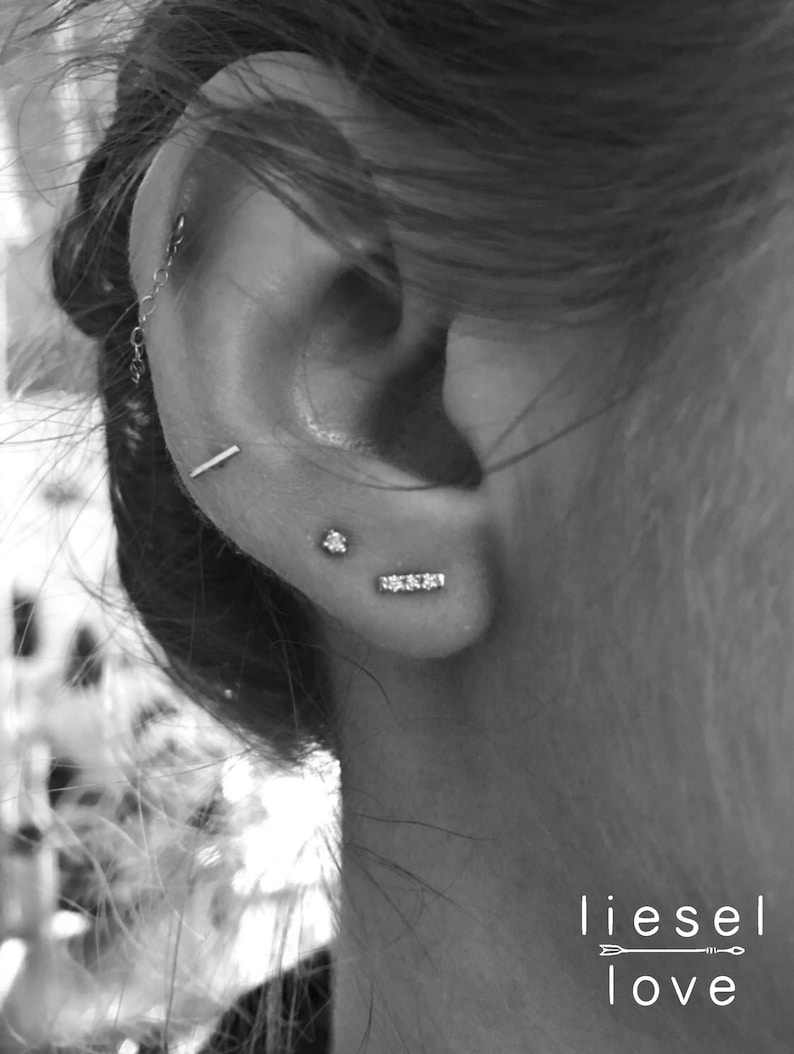 May include: A black and white image showing a set of silver-toned earrings worn in an ear. The jewelry includes a small stud, a thin horizontal bar, a three-stone bar, and a chain-linked piece. The text "liesel love" appears in the bottom corner.