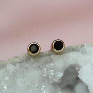 May include: A pair of stud earrings featuring round, dark black stones set in polished gold-toned metal bezels. The earrings are circular in shape with a smooth, metallic finish surrounding the dark center, creating a simple and classic design.