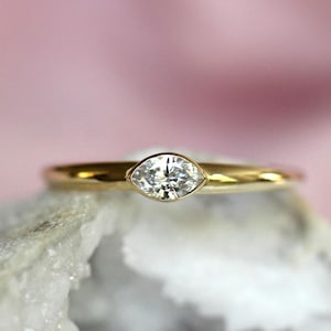 May include: A gold ring featuring a single marquise-cut clear gemstone set horizontally in a bezel setting. The band is smooth and polished, showcasing a clean, minimalist design that highlights the elongated shape of the central stone.