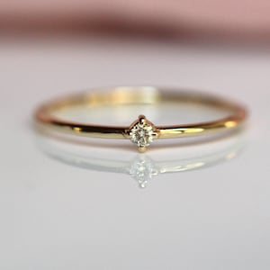 May include: A thin gold band ring featuring a single, small, round, clear faceted gemstone set in a four-prong setting at the centre. The metal has a polished, reflective finish, and the ring is positioned horizontally, showing its circular shape.