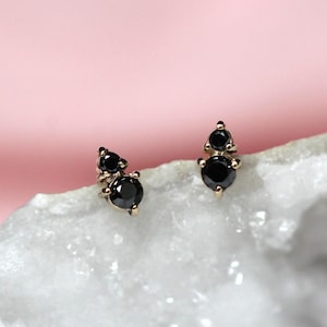 Puede incluir: Un par de pendientes de botón con dos piedras negras redondas engastadas en metal dorado. Cada pendiente consta de una piedra más pequeña situada sobre una más grande, creando un diseño de racimo vertical sobre una superficie de cristal.