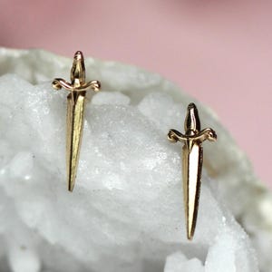 May include: A pair of gold-toned stud earrings shaped like miniature daggers. Each earring features a pointed blade, a central crossguard, and a detailed handle. The metallic finish reflects light, highlighting the sharp, symmetrical design of the blades.