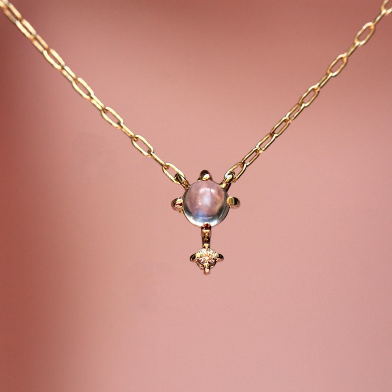 May include: A delicate gold necklace with a moonstone pendant. The pendant features a round moonstone surrounded by small gold accents and a smaller diamond below.