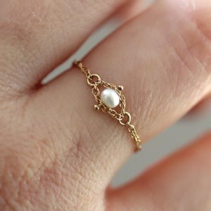 Puede incluir: Un delicado anillo de cadena de oro con una pequeña perla blanca de forma ovalada centrada en un marco de metal decorativo. El anillo está construido con finos eslabones de oro entrelazados que forman una banda flexible con acabado pulido.