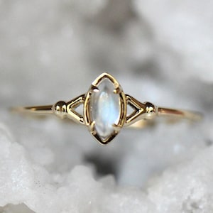 May include: A gold ring featuring a marquise-cut moonstone set in a prong setting. The band has a delicate design with small metal spheres on either side of the central stone, creating a symmetrical look with a polished finish and iridescent gem.
