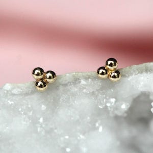 May include: A pair of gold stud earrings, each featuring three small, polished spherical beads arranged in a triangular cluster. The earrings have a smooth, reflective metallic finish and are designed as simple, compact geometric shapes.