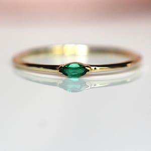 May include: A thin gold band ring featuring a single marquise-cut green gemstone set horizontally in the center. The metal has a polished finish, and the gemstone is a vibrant, deep emerald green color, creating a simple and refined appearance.