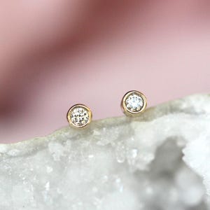 May include: A pair of stud earrings featuring round, clear, faceted stones set in polished gold-toned metal bezels. The circular settings hold the stones securely, creating a simple and reflective design that catches the light on each earring.