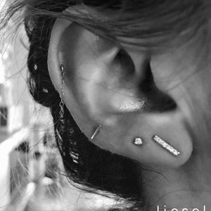 May include: A close-up of a person's ear with multiple piercings. The ear is pierced with a small chain, a stud, and a bar earring. The image is in black and white and has the text "liesel love" at the bottom.