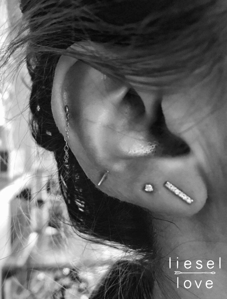 May include: A close-up of a person's ear with multiple piercings. The ear is pierced with a small chain, a stud, and a bar earring. The image is in black and white and has the text "liesel love" at the bottom.