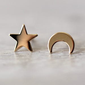 May include: A pair of gold-toned stud earrings featuring a five-pointed star shape and a crescent moon shape. Both earrings have a smooth, polished metallic finish and are designed as individual pieces to be worn as a matching set of celestial symbols.