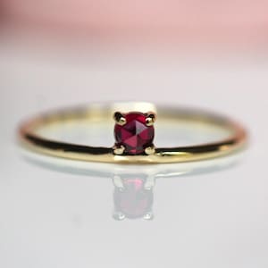 May include: A thin gold band ring featuring a single, small, round-cut deep red gemstone set in a four-prong setting. The ring has a polished metallic finish and is positioned horizontally, displaying a clear reflection on the surface below.