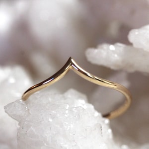 Peut inclure: Une bague en or poli avec un design en forme de V ou chevron. L'anneau présente une surface métallique lisse et réfléchissante qui se courbe vers un point central, créant une silhouette angulaire distincte posée sur une structure cristalline.