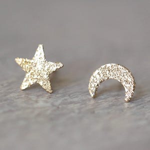 May include: A pair of gold-toned stud earrings featuring a five-pointed star shape and a crescent moon shape. Both earrings have a distinct, rough, and textured surface finish that catches the light, creating a sparkling appearance on each piece.