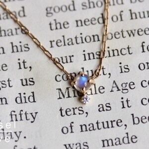 May include: A delicate gold chain necklace featuring a small, iridescent moonstone pendant with a tiny diamond accent. The necklace is shown on a page of a book with text.