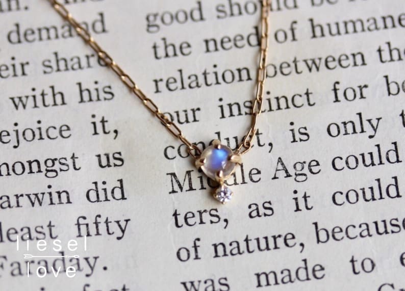 May include: A delicate gold chain necklace featuring a small, iridescent moonstone pendant with a tiny diamond accent. The necklace is shown on a page of a book with text.