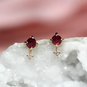 以下が含まれることがあります： 小さな赤い宝石と、その下にぶら下がっている単一のダイヤモンドが付いた、金色のイヤリングのペア。イヤリングは白い背景に置かれています。