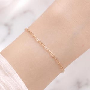 Puede incluir: Una delicada pulsera de cadena en tono dorado con eslabones rectangulares alargados estilo clip. La pulsera se lleva alrededor de la muñeca, mostrando un diseño sencillo con un acabado metálico pulido y un cierre sutil y discreto.