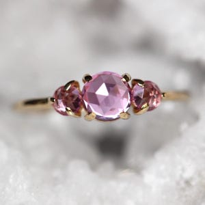 Puede incluir: Un anillo de oro con una piedra preciosa rosa redonda y facetada en el centro, flanqueada por dos piedras rosas más pequeñas. Cada piedra está sujeta por cuatro garras de oro, creando un diseño de tres piedras sobre una banda de oro pulido.