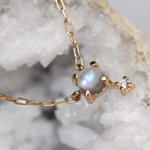 May include: A delicate gold chain necklace featuring a round moonstone pendant with a small diamond accent. The pendant is set in a gold claw setting.