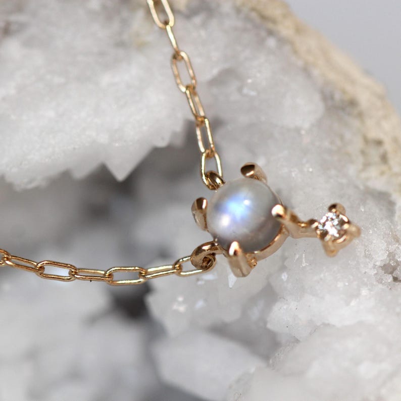 May include: A delicate gold chain necklace featuring a round moonstone pendant with a small diamond accent. The pendant is set in a gold claw setting.