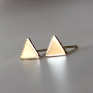 May include: A pair of gold-toned stud earrings featuring a simple, polished triangle shape. Each earring has a smooth, reflective surface and a thin metal post extending from the back, designed for secure placement in a pierced earlobe.