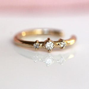 May include: A gold ring featuring an open band design with three clear, round-cut stones set in prongs along the top. The central stone is slightly larger than the two flanking stones, creating a balanced arrangement on the polished gold metal band.