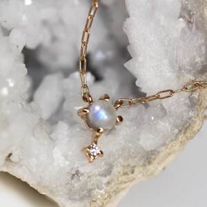 May include: A delicate gold chain necklace featuring a moonstone pendant with a small diamond accent. The pendant is set in a gold setting and has a subtle iridescent shimmer.