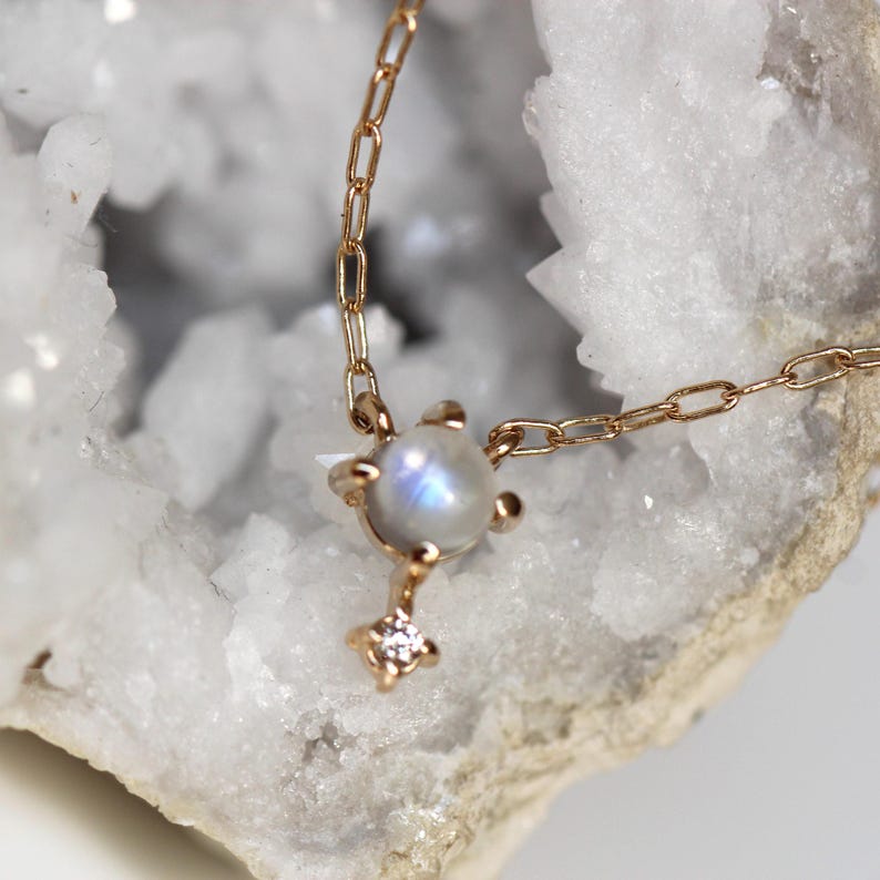May include: A delicate gold chain necklace featuring a moonstone pendant with a small diamond accent. The pendant is set in a gold setting and has a subtle iridescent shimmer.