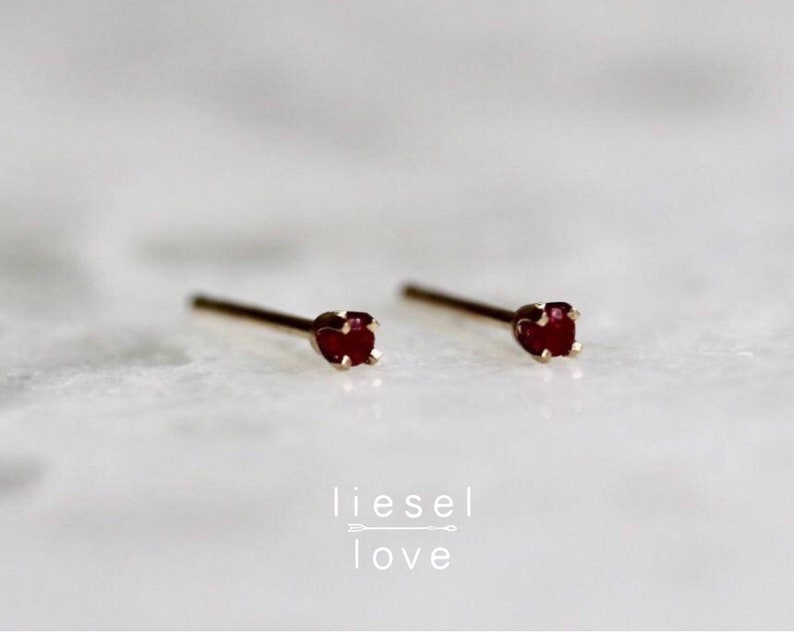 May include: A pair of small, gold stud earrings with a single, dark red gemstone in each.