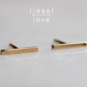 May include: A pair of gold-toned metal bar stud earrings with a smooth, polished finish and a post backing. The text "liesel love" with an arrow graphic is positioned above the earrings in a clean, white sans-serif font.