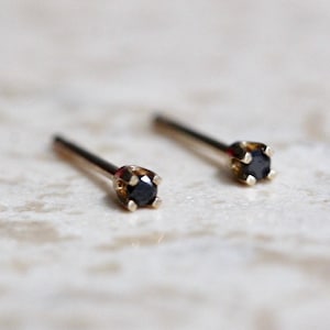 May include: A pair of stud earrings featuring small, round, black stones set in four-prong gold metal settings. Each earring has a thin, straight post extending from the back of the setting, designed for insertion through a pierced earlobe.