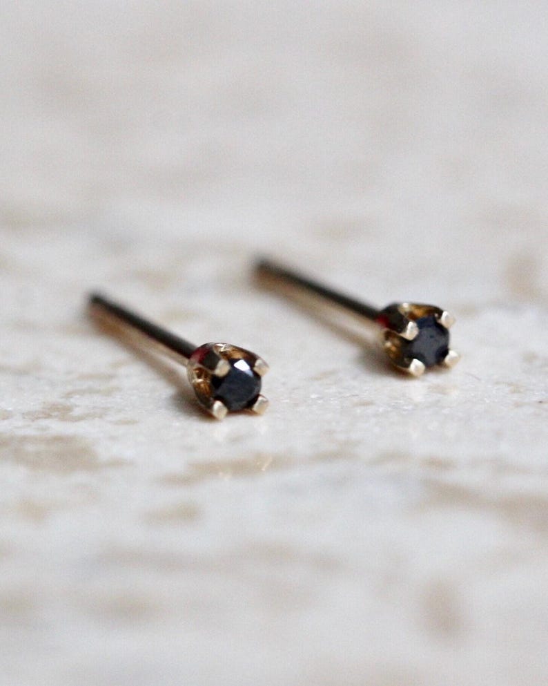 May include: A pair of stud earrings featuring small, round, black stones set in four-prong gold metal settings. Each earring has a thin, straight post extending from the back of the setting, designed for insertion through a pierced earlobe.
