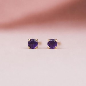 May include: A pair of stud earrings featuring round, deep purple amethyst gemstones set in four-prong gold settings. The stones are faceted to catch the light, and the metal prongs securely hold each circular gem in place, creating a classic design.