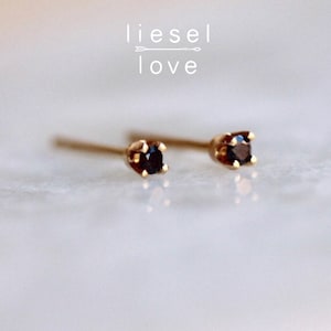 May include: A pair of gold stud earrings featuring small, round, dark black gemstones held in four-prong settings. The text "liesel love" with an arrow graphic is positioned above the earrings. The earrings have thin posts extending backward.