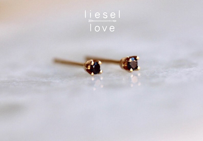 May include: A pair of gold stud earrings featuring small, round, dark black gemstones held in four-prong settings. The text "liesel love" with an arrow graphic is positioned above the earrings. The earrings have thin posts extending backward.