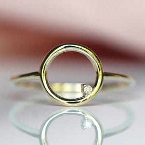 May include: A gold ring featuring a circular open frame design. A single small, clear, round-cut gemstone is set into the lower right side of the circular band. The metal has a polished, reflective finish that catches the light.