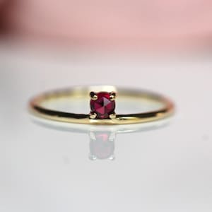 May include: A delicate gold band ring with a single, small, round, red gemstone set in the center.